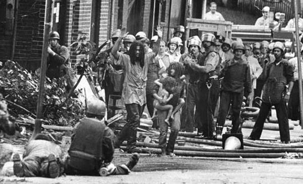 Después del feroz tiroteo e inundación causada por los policías, miembros de MOVE, incluyendo niños pequeños, emergen de cara a las armas de fuego con que los policías los están apuntando.