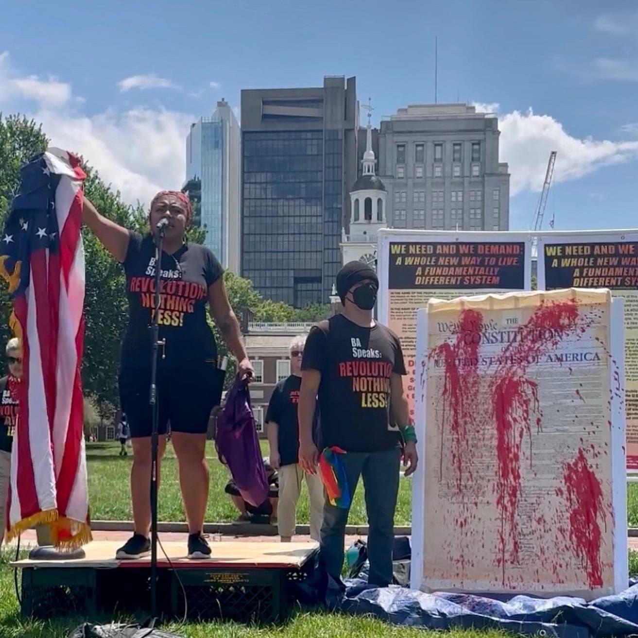 Revcoms holding the U.S. flag and red-paint-splattered constitution, July 4, in front of Independence Hall, Philadelphia.