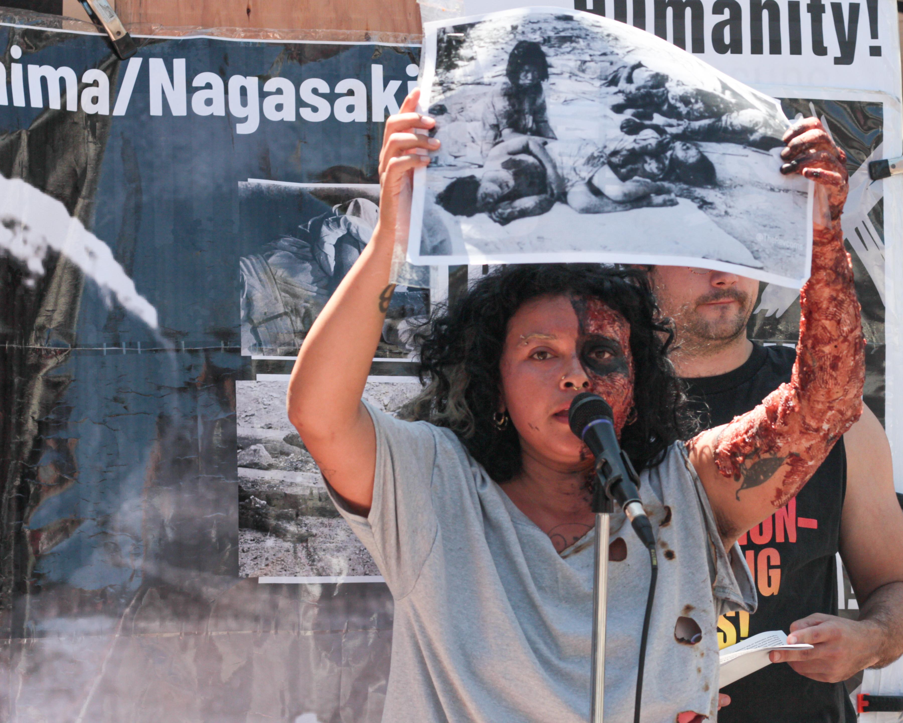 Woman whose face is disfigured by makeup holding a large photo of Hiroshima bombing.