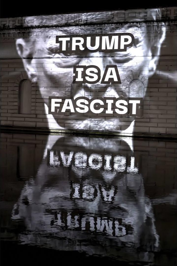 Trump's face in lightshow on wall with "Trump is a Fascist" written on it.