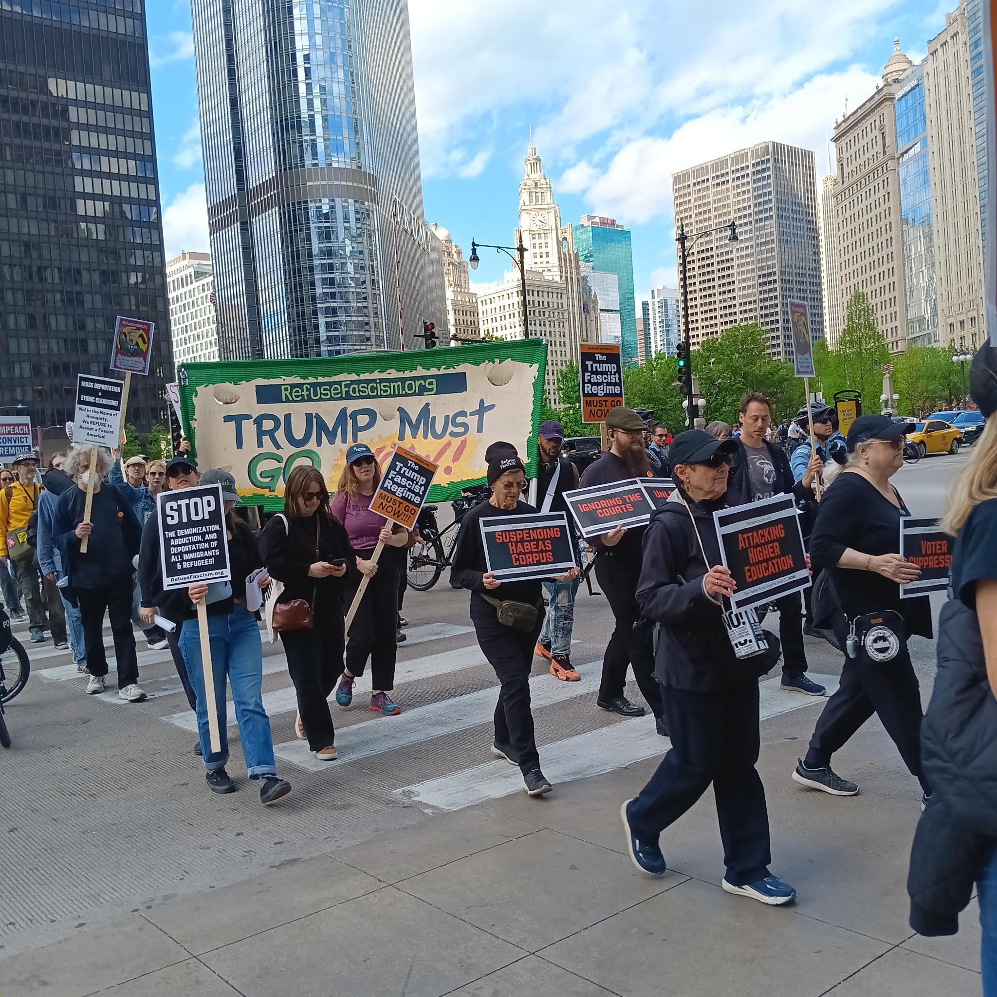At the Chicago, Refuse Fascism rally signs with horrors of Trump's fascist directives march in front of Trump Must Go Now banner.