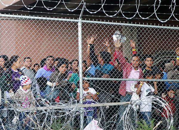 Immigrants crowded behind a fence under a bridge.